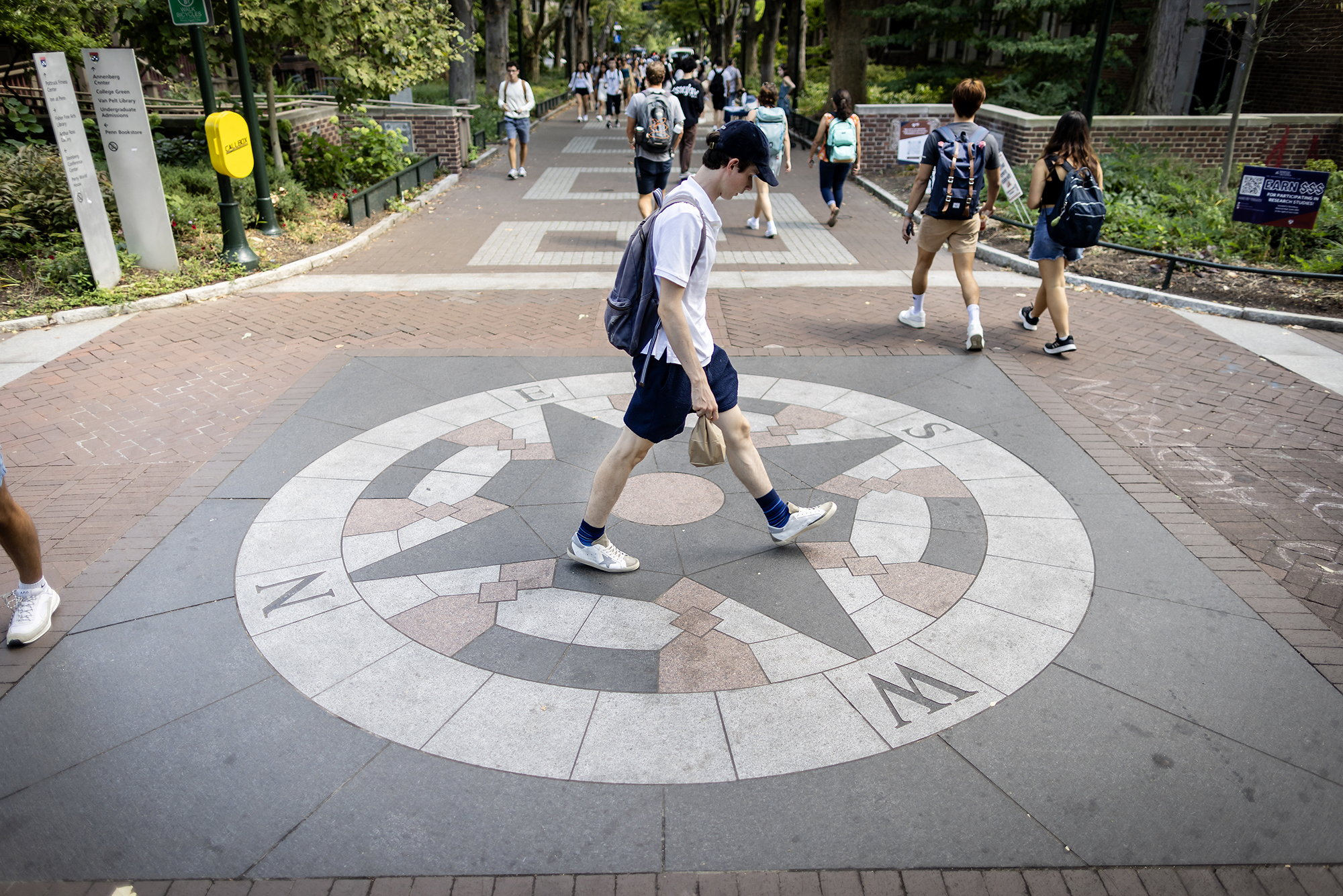 A person in shorts and a baseball hat wearing a backpack walks across a compass embedded in Locust Walk on Penn campus.
