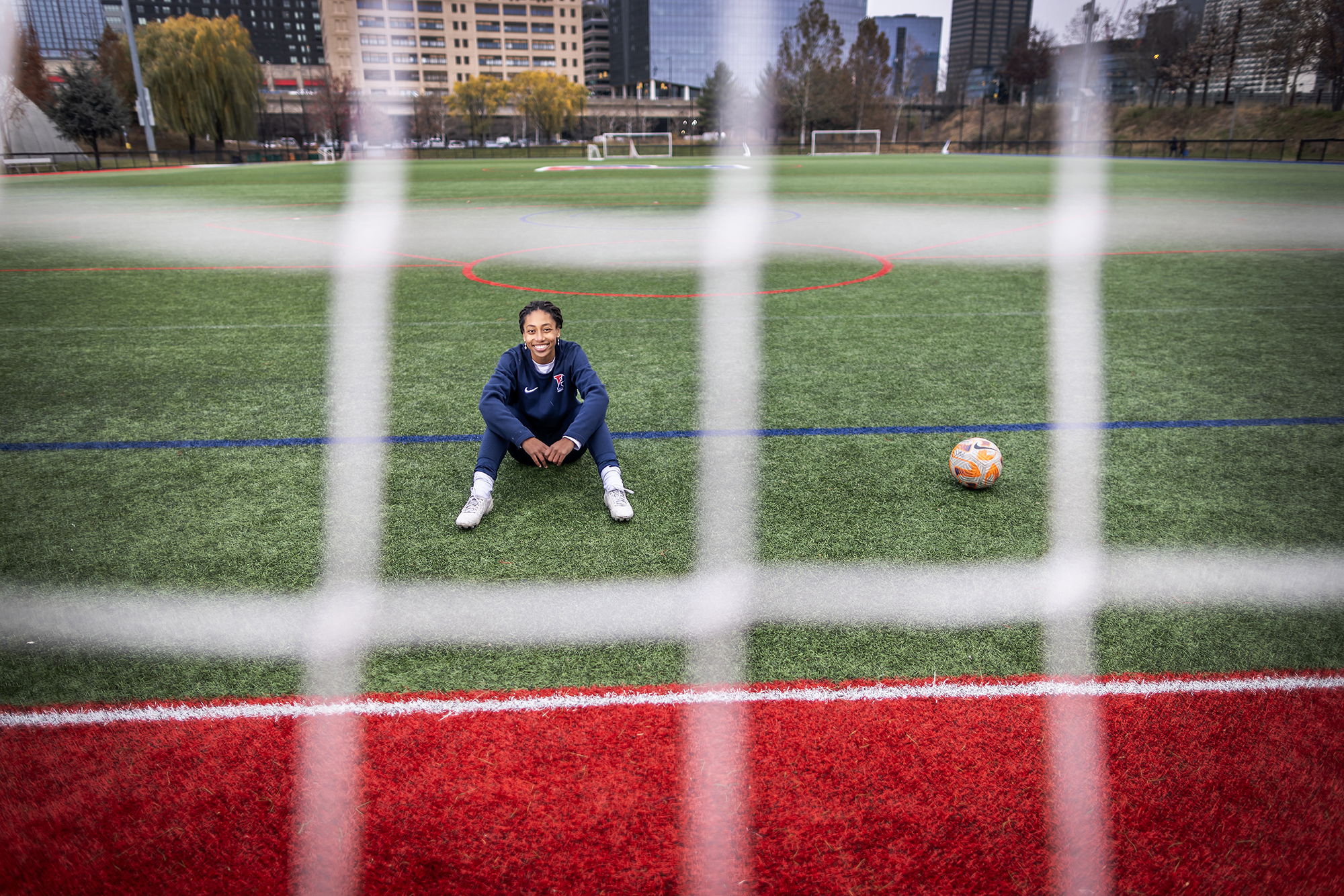 Ginger Fontenot, wearing a Penn sweatsuit, sits on the field at Penn Park beside a soccer ball.