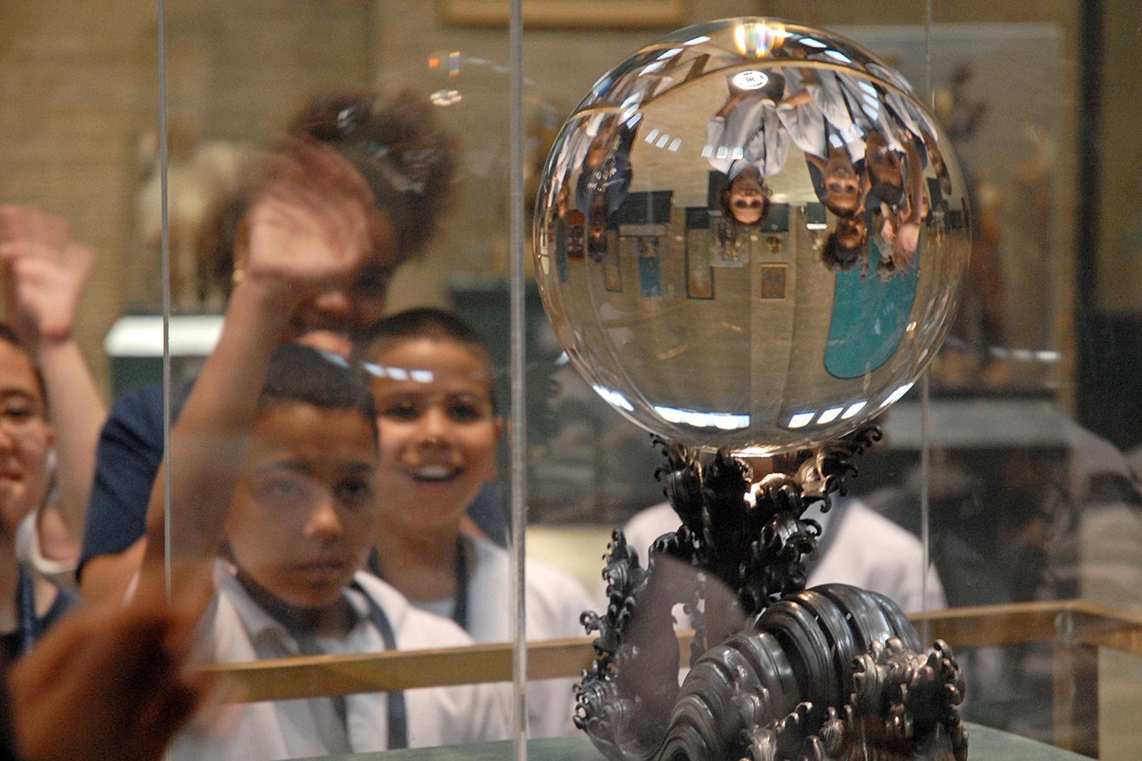 A group of school children in white polo shirts wave and gesture at the crystal ball, seeing themselves reflected upside down 