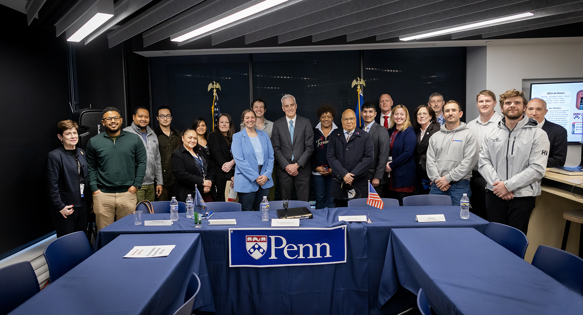 Secretary of Veterans Affairs smiles for a photo with students and staff from Penn, at table with Penn logo in front