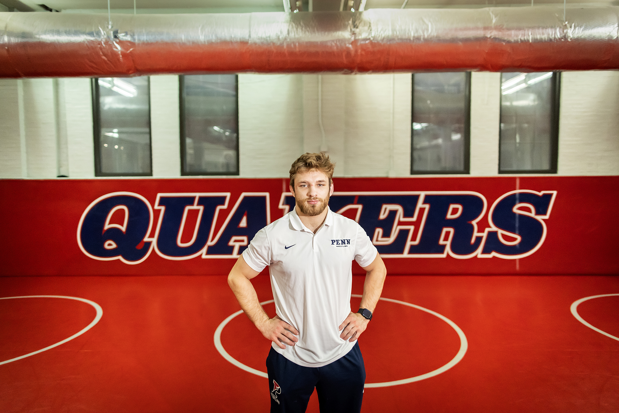 Wrestling Adam Thomson stands in the wrestling room at Penn, hands on hips, with a sign reading Quakers behind him.