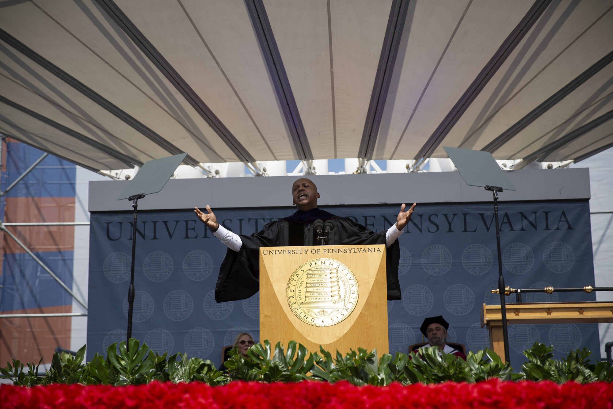 Bryan Stevenson reaches his arms out wide while speaking at a podium on stage at commencement.