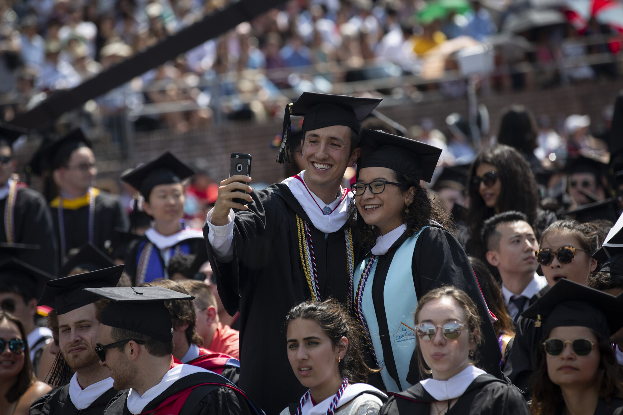 Two graduating students stand and take a selfie on the field during commencement