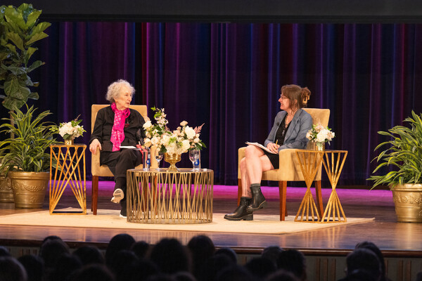 Margaret Atwood and Emily Wilson sitting in chairs on a stage with flowers on a table in front of them and tall green plants to the side