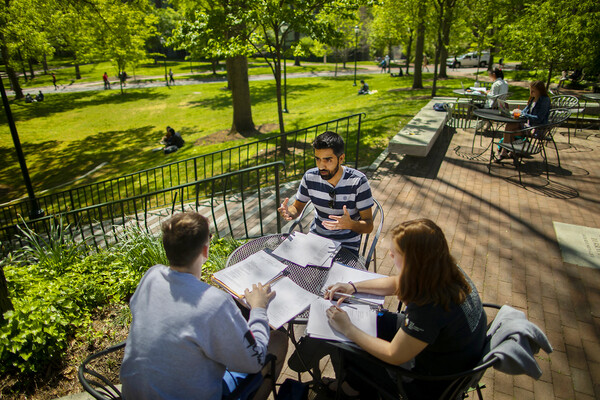 Three people sitting at a small, round table outside, with greenery in the background.