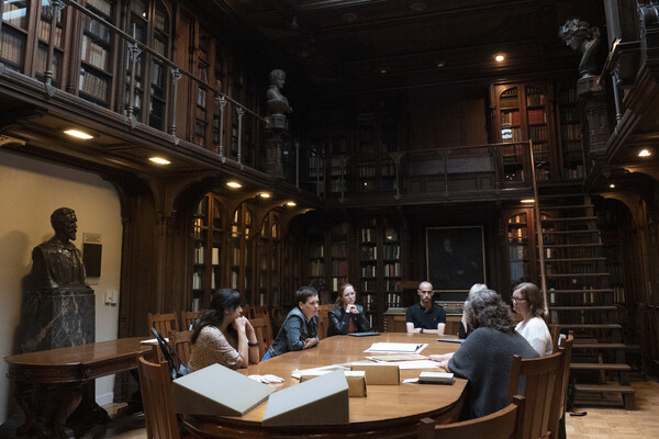 A group of people sitting around a rectangular wooden table on the bottom floor of a two-story room in a library adorned with books and busts.