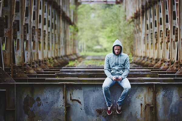 Person wearing a hoodie sits on a structural beam alone in an isolated area holding a smartphone.