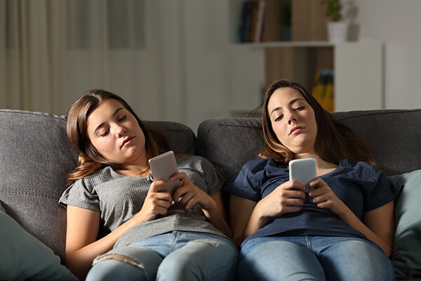 Two teens lay on couch with smartphones looking bored