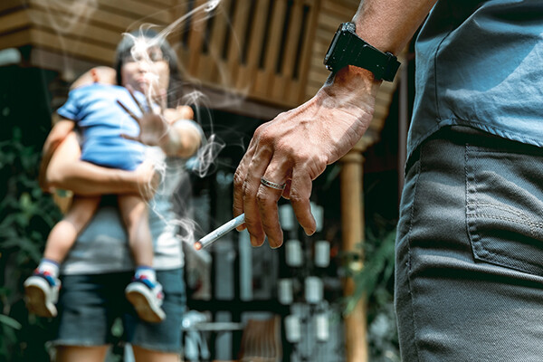 hand holding a lit cigarette in foreground, parent holding a toddler and putting out their hand to indicate stop smoking in background.