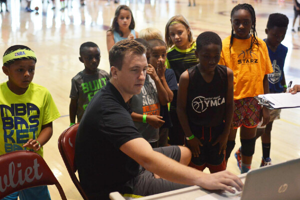 a person looking at a laptop surrounded by kids watching the screen standing in a basketball court