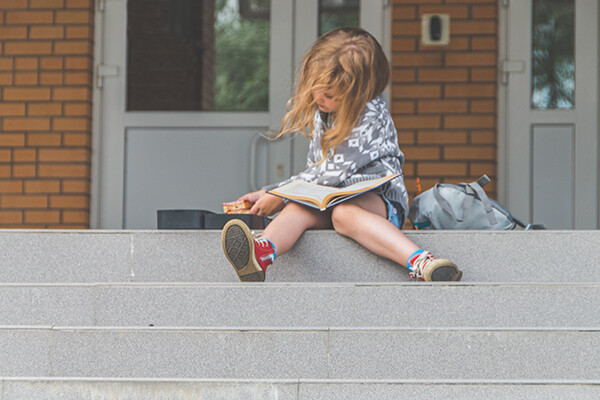 Young child sits on front steps with a book in their lap taking a sandwich out of a lunchbox.