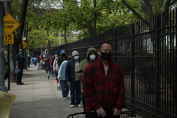 A line of people outside a food bank wait on a sidewalk practicing social distancing and wearing face masks.