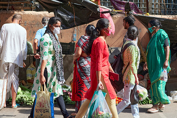 Several people wearing face masks walk through an outdoor market carrying bags of groceries in India