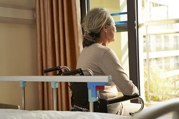 Elderly person in a wheelchair sits by an open window next to their bed in a nursing home.