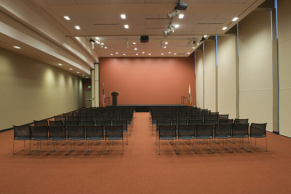A conference room full of empty chairs in front of an empty podium on a stage with an American flag on the left side.
