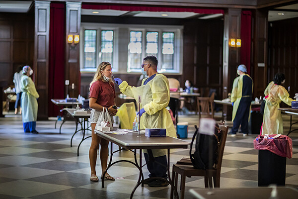 Two people standing by a table in Houston Hall, one wears PPE and administers a nasal swab to a student.