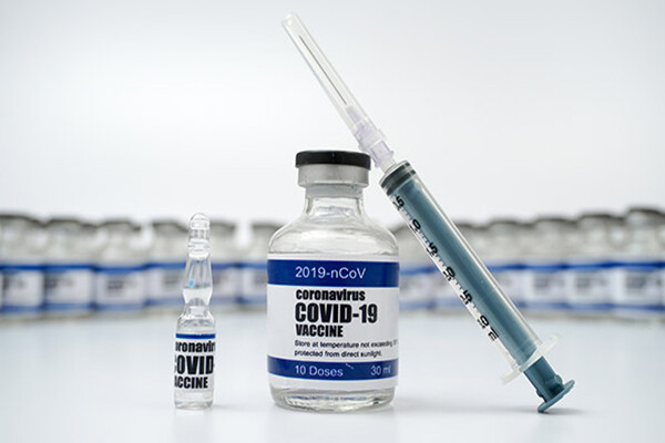 Bottle of liquid COVID-19 vaccination with a syringe lying against it in front of a row of vaccine bottles in the background.
