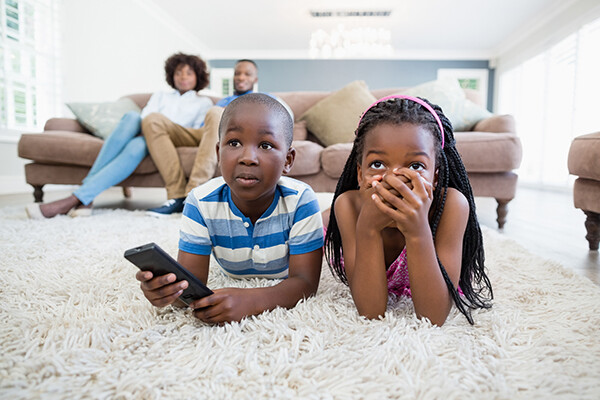 Black family watching television, two children lying on the carpet, two parents on the couch behind them.