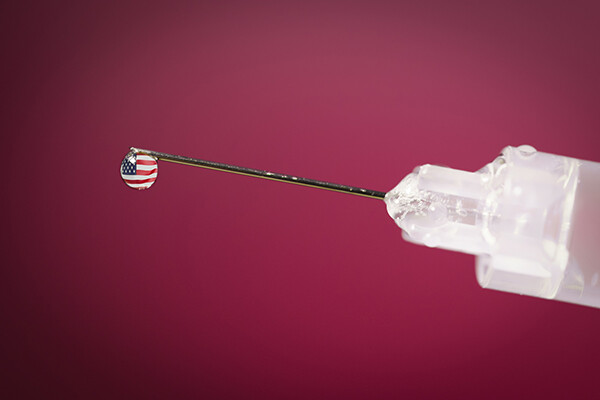 Closeup of a syringe with a droplet at the tip of the needle reflecting the American flag pattern.