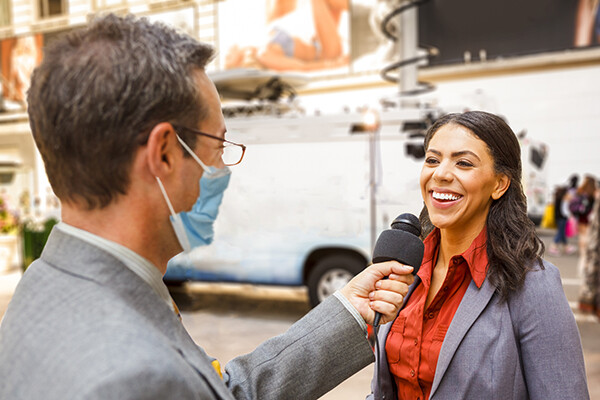 Masked news reporter holds a microphone to an interviewee laughing and responding.