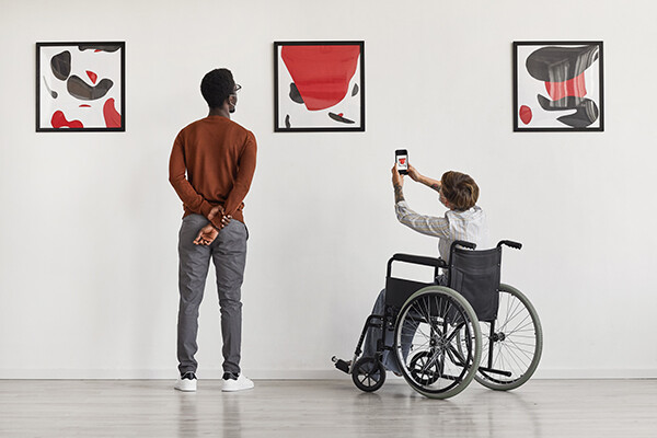 Two people look at three paintings on a wall in a museum, one is in a wheelchair taking a photo with a smartphone.