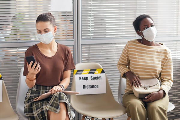 Two people, one Black one white, sitting in a waiting room wearing masks.
