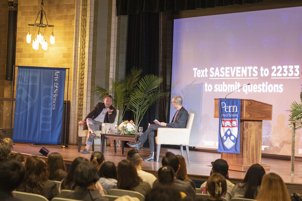 Two men sit on a stage in front of a crowd. A screen in the back says, "Text SASEVENTS to 2233 to submit questions"
