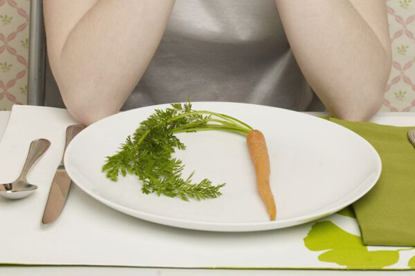 A person seated at a dining table with their elbows on the table, with one single carrot on the plate before them.