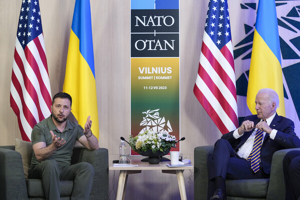U.S. President Joe Biden and Ukraine’s President Volodymyr Zelenskyy are seated next to each other, separated by a side table with flowers and in front to the American and Ukrainian flags on each side of them. Behind them is a sign reading NATO/OTAN Vilnius Summit/Sommet.