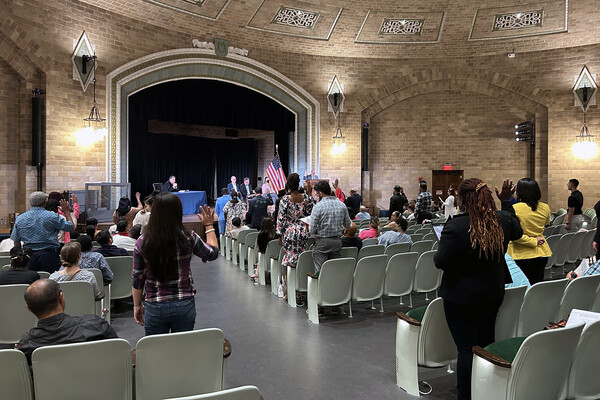 Newly naturalized U.S. citizens raise their right hands to take the oath of citizenship in the Harrison Auditorium at the Penn Museum, with officials on a stage in front of them and an American flag on the right side of the stage.
