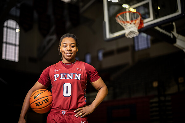 Jordan Obi, wearing her red Penn jersey, stands with her back to the basketball with a ball under her right arm.