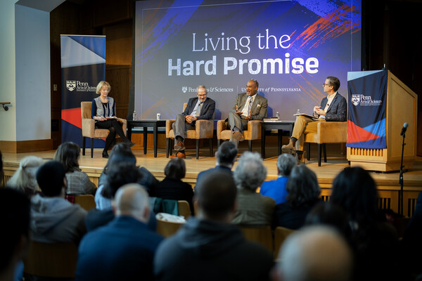 Four people sit in chairs on an auditorium stage in front of a sign reading Living the Hard Promise.