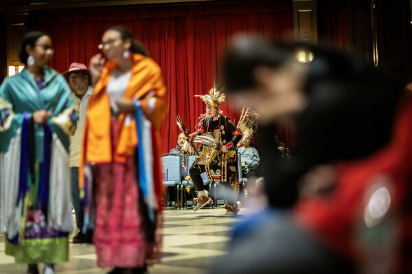 In the foreground, female dancers with shawls. In the background, a young man in warrior regalia. 