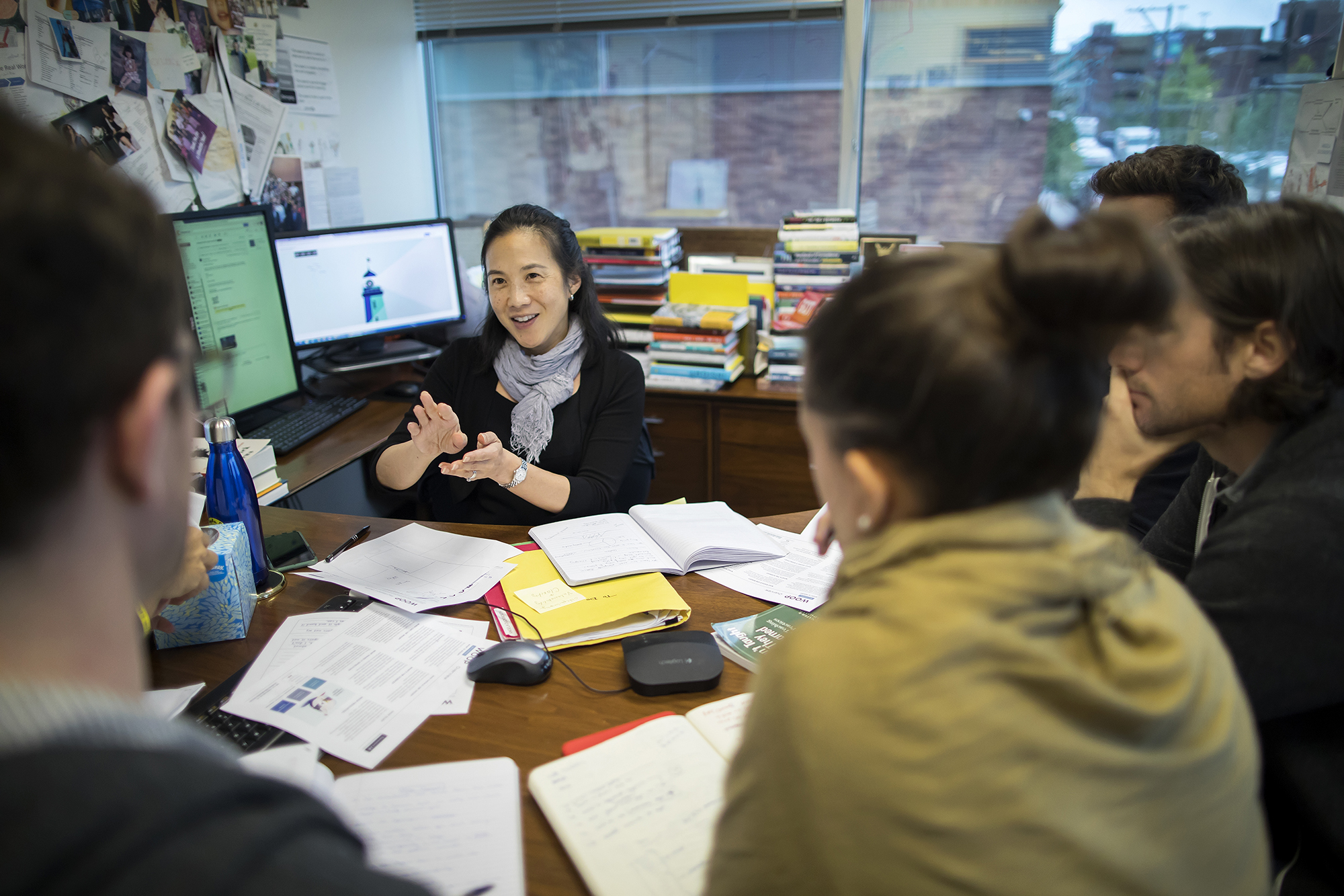 A person sitting at a desk covered in papers, with a computer screen in the background. Four people are blurry, in the foreground. They are all engaged in conversation.