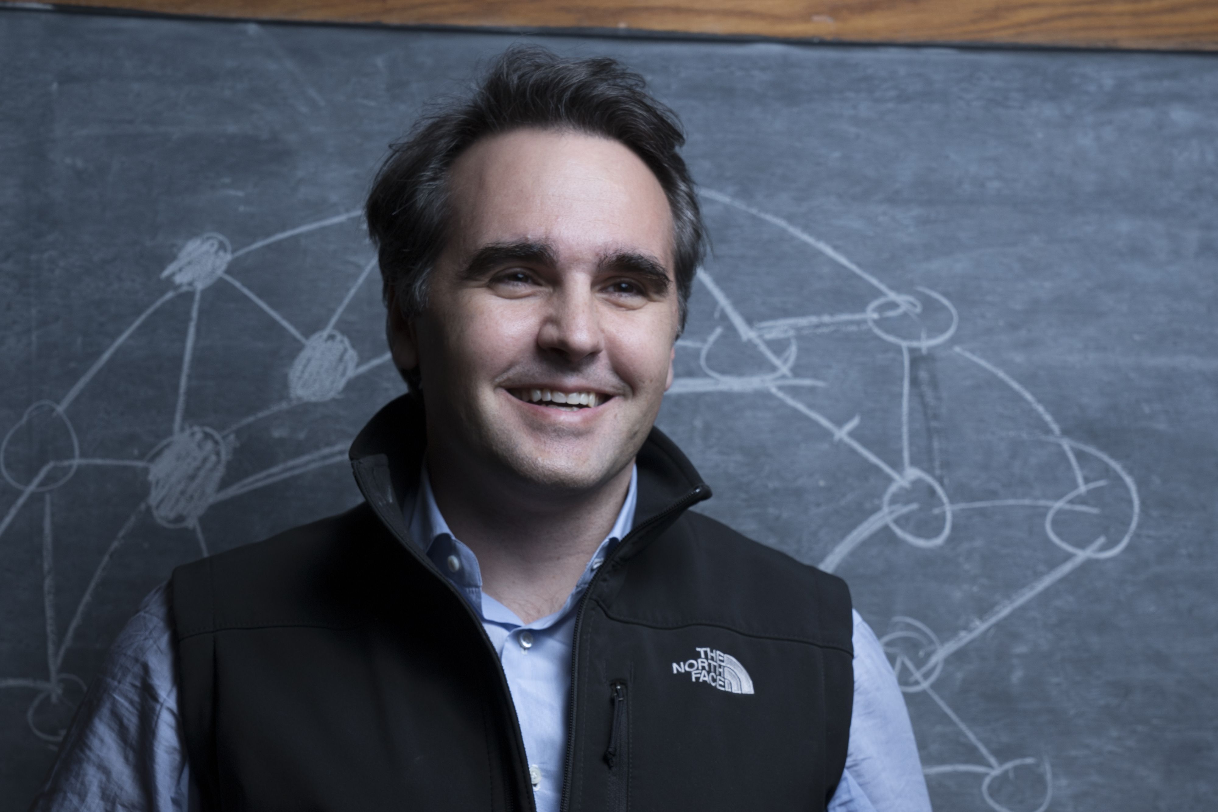A smiling person in a blue button down shirt with a black "The North Face" vest over it standing in front of a chalkboard with lines and circles, some of which are filled in.