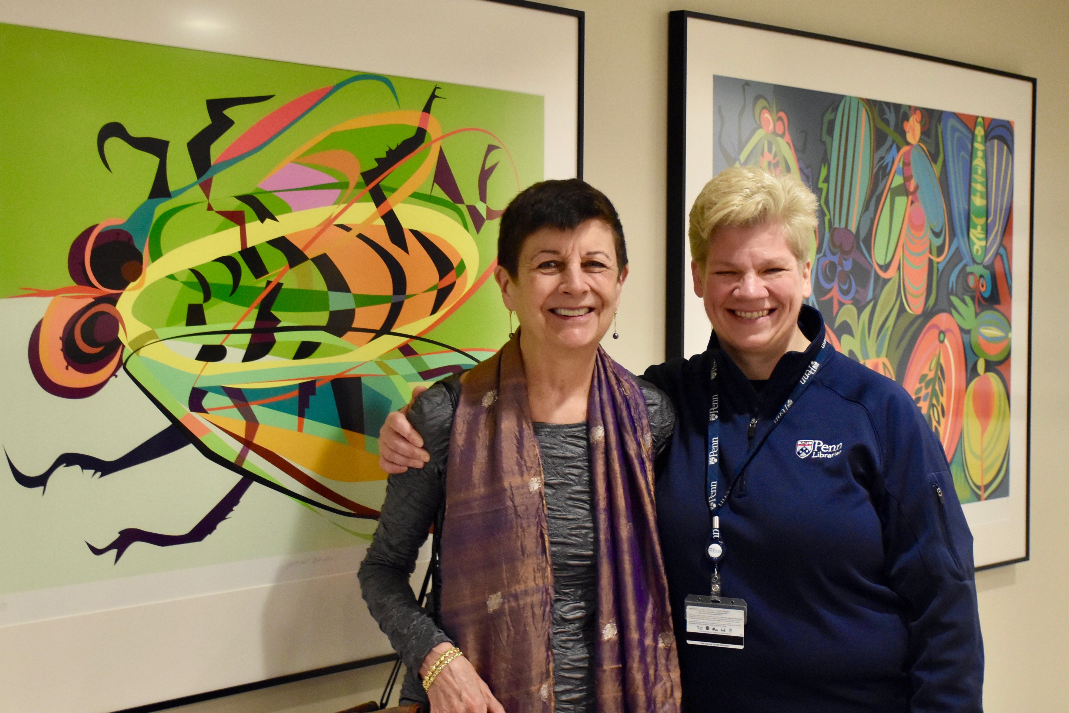 Penn-Libraries-Regan-Kladstrup-standing-with-alumna-Caroline-Schimmel-in-front-of-brightly-colored-prints-by-woman-Ecuadorian-artist-depicting-rainforest
