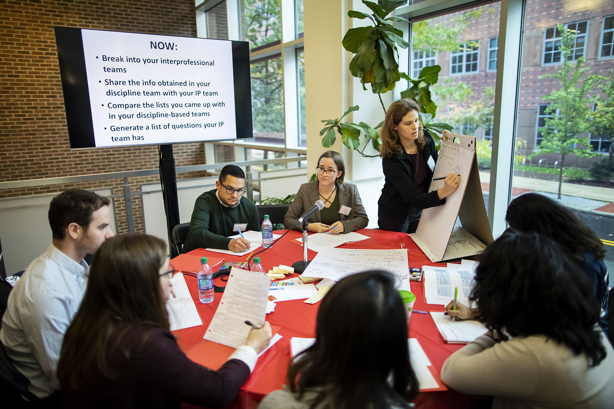 Students sit around a table as a faculty member takes notes on an easel, a screen with instructions is up in the background