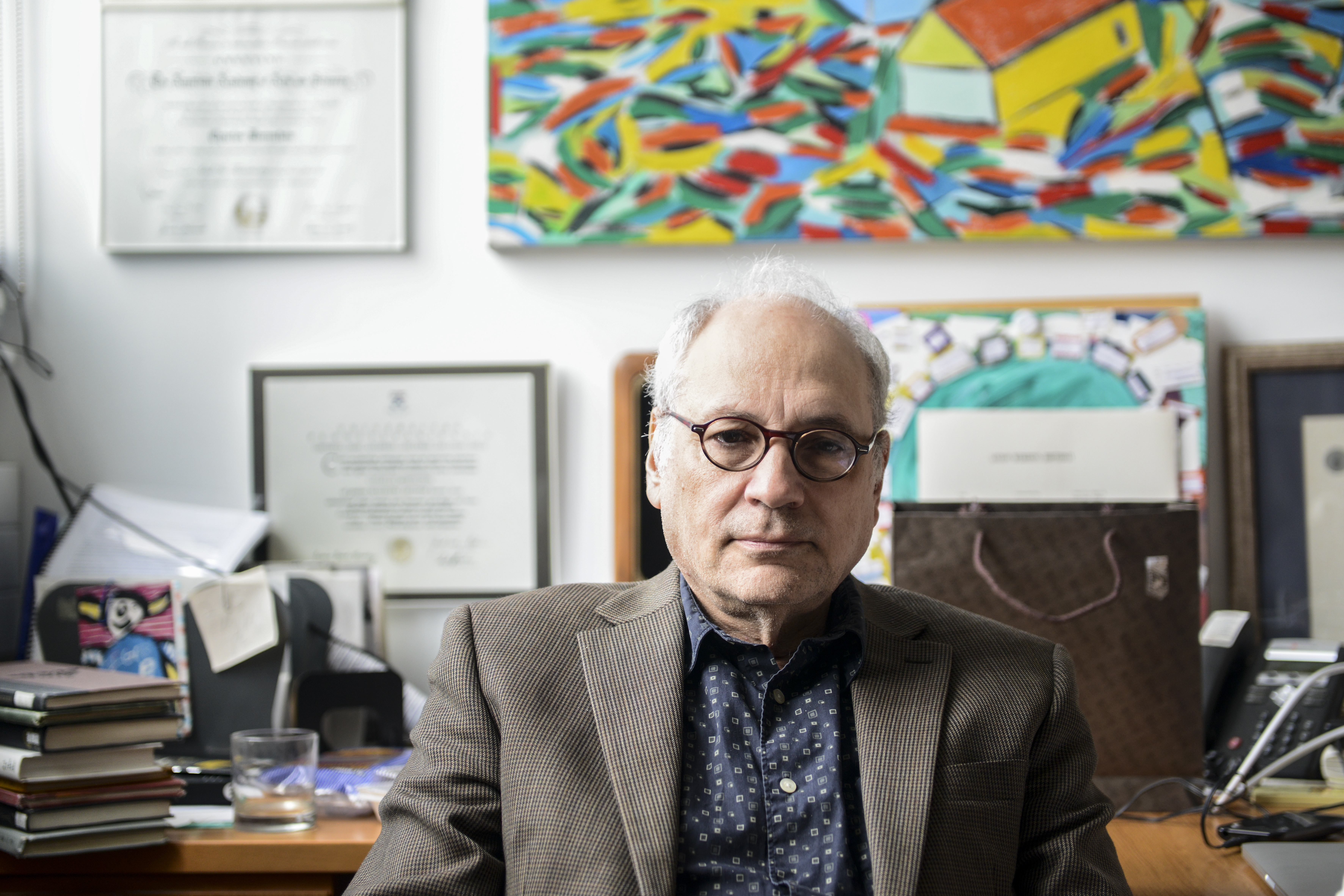Charles Bernstein at his desk in his office with a colorful painting by his wife on the wall behind him