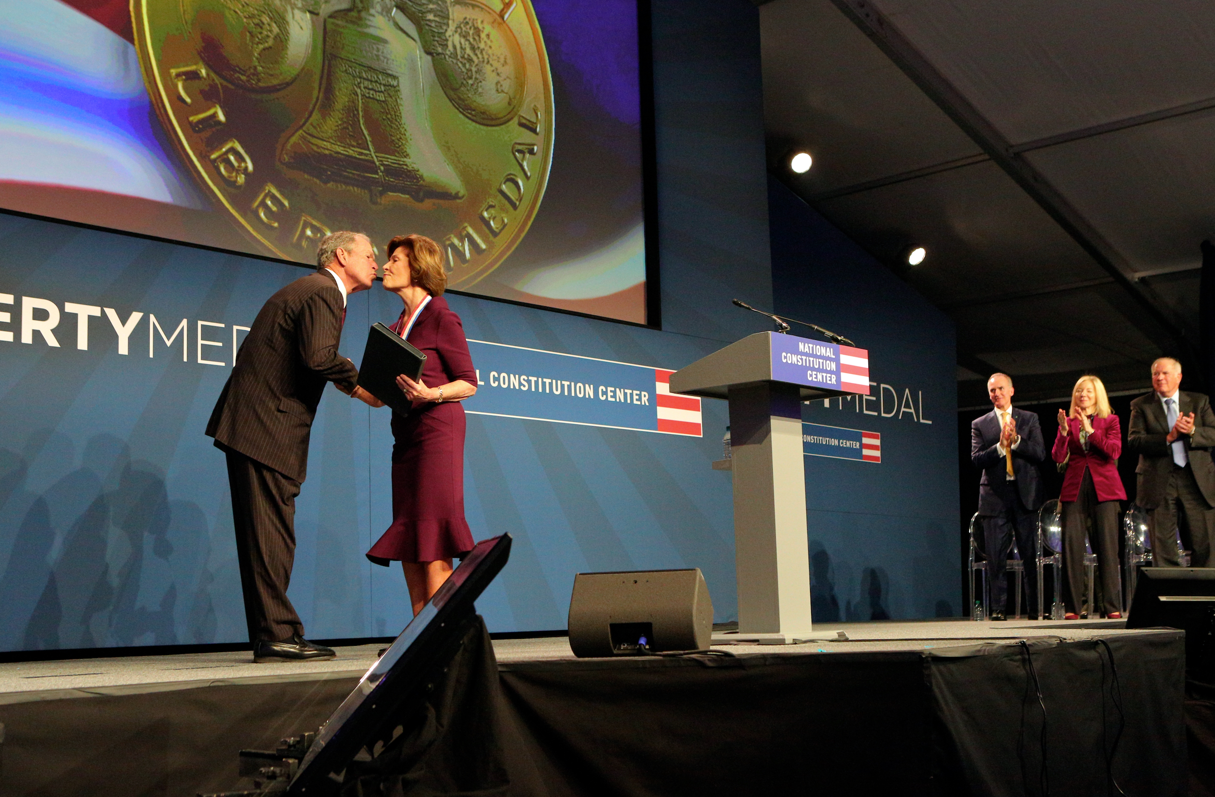 George W. Bush accepting award from Laura Bush on stage with President Gutmann clapping at the far end of the stage