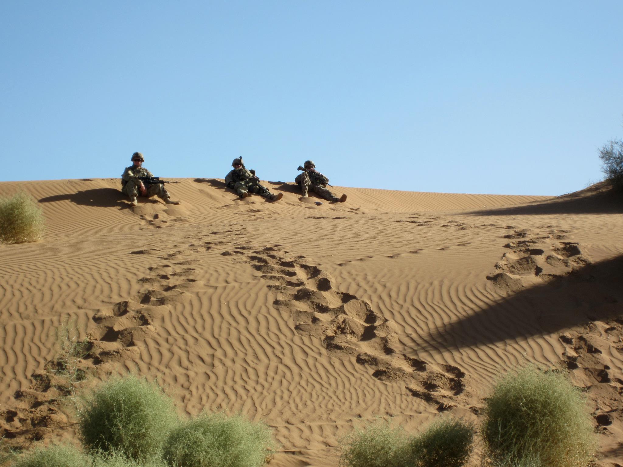 Three Army soldiers sit on top of a hill in a desert. 