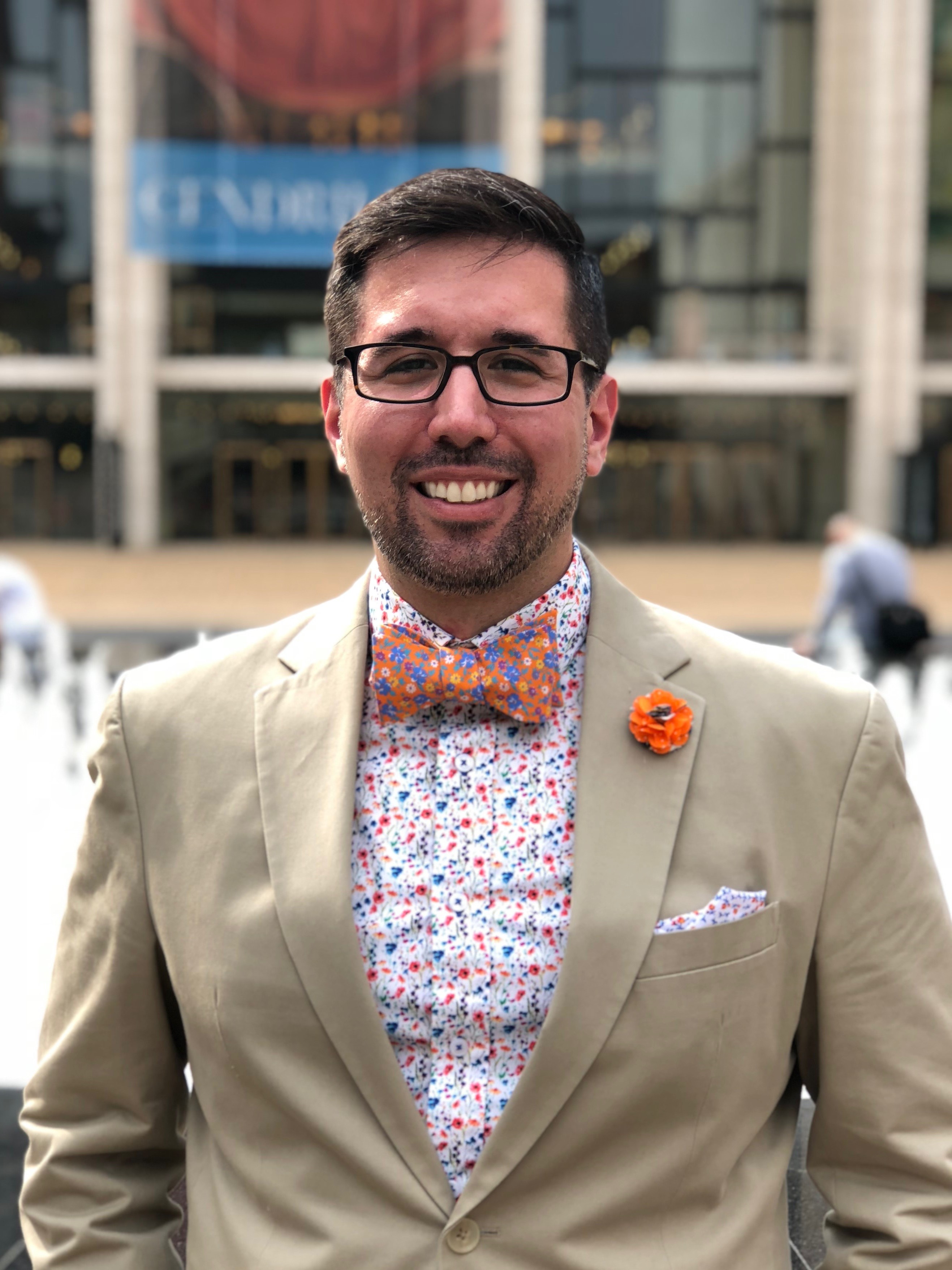 Mark Lo poses wearing a floral shirt and tie and a tan jacket.