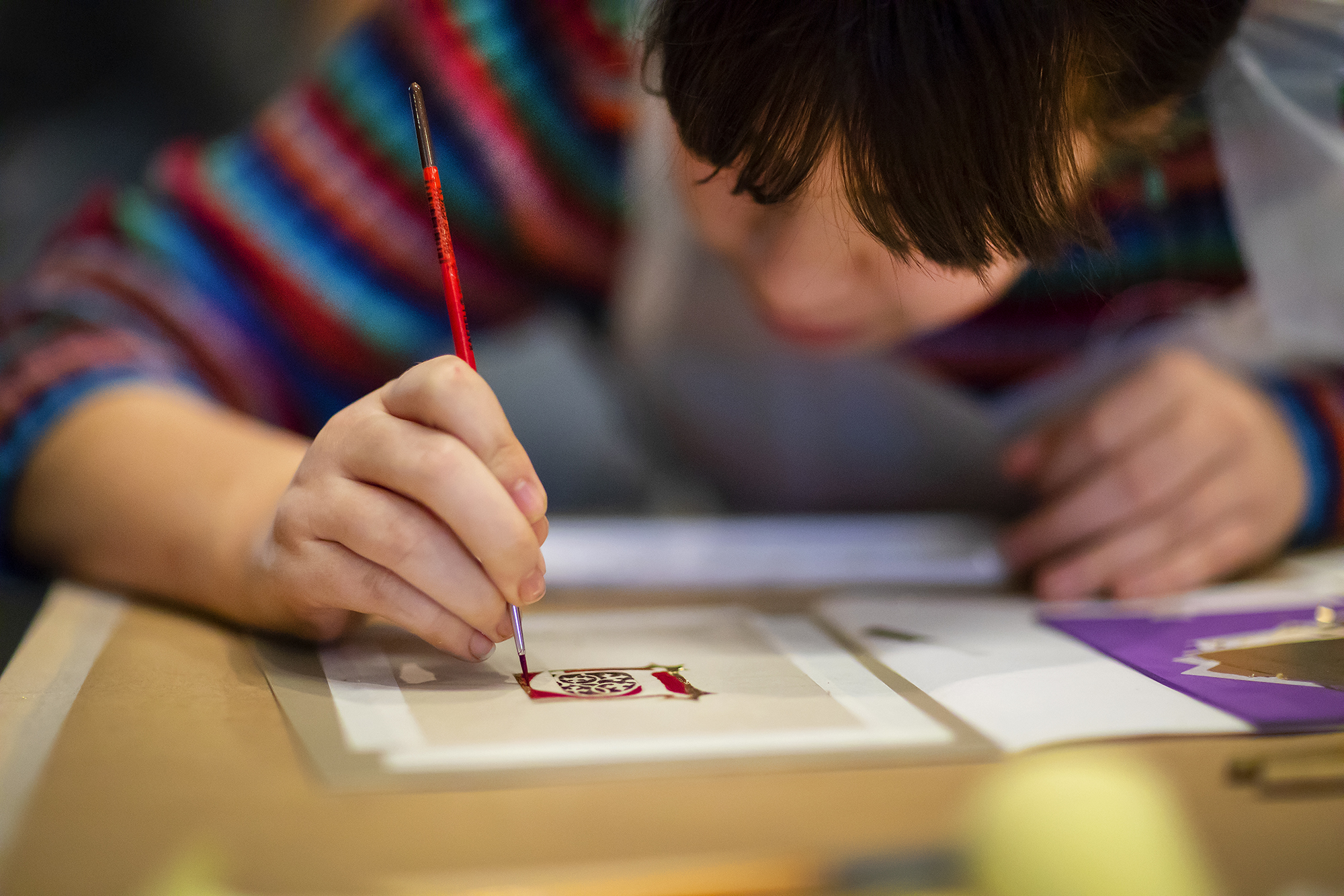 closeup of student with paint brush painting red paint on small image on paper at a table