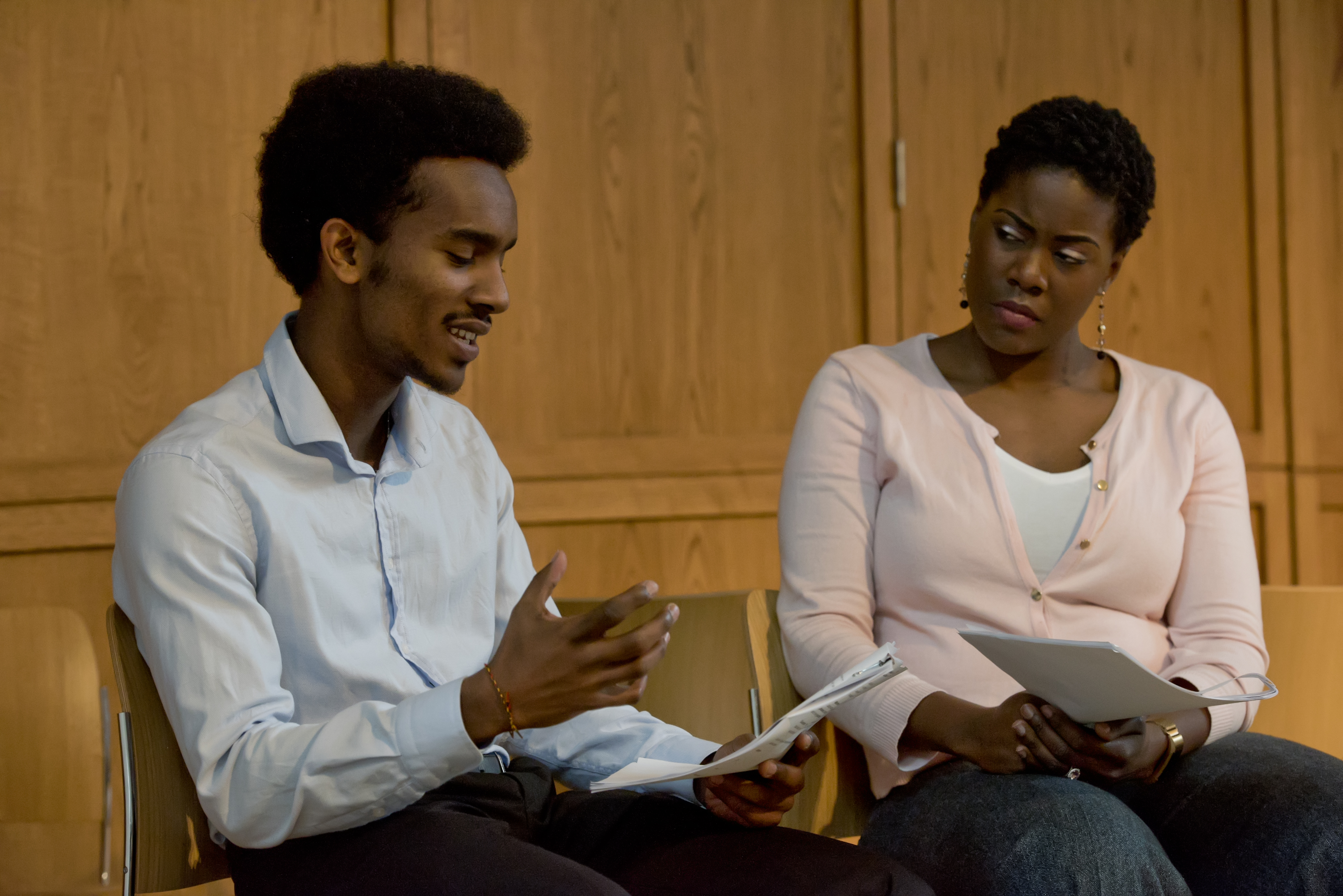 two people seated with one reading aloud from a script while the other listens intently