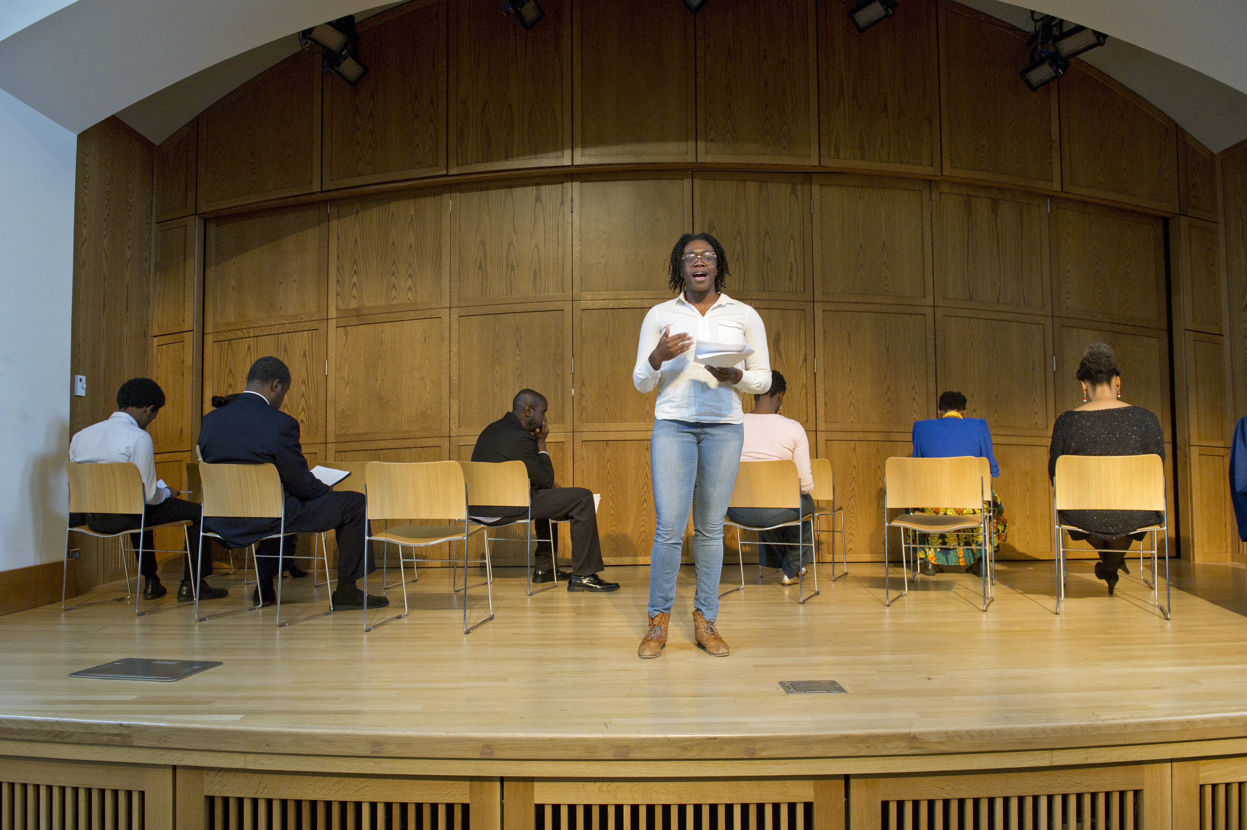 performer on stage reading from a script at the Community Monologue event with seated performers behind her on stage