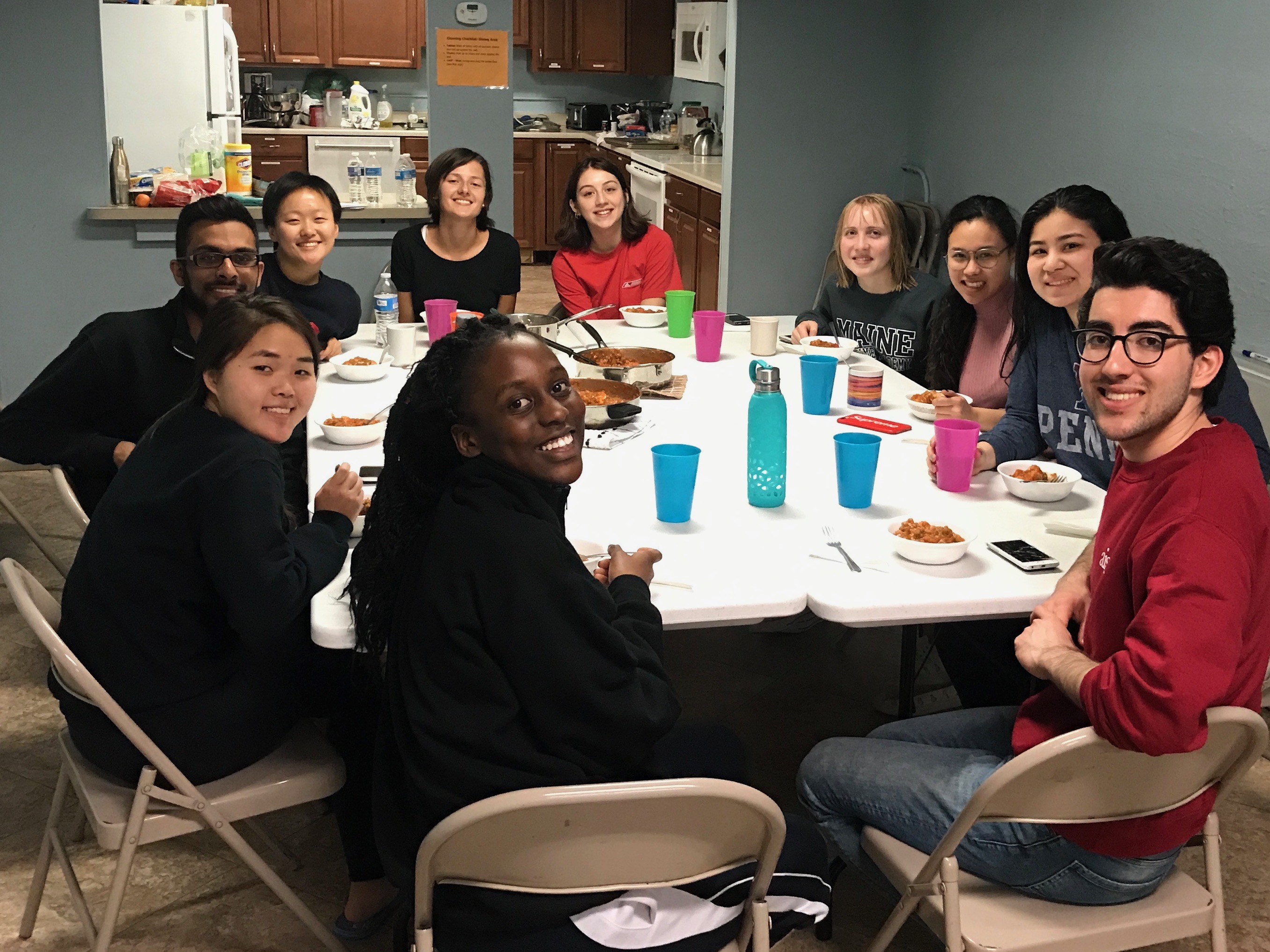 Ten students around a table all looking and smiling at the camera. The table has bowls of food and cups and is next to a kitchen. 