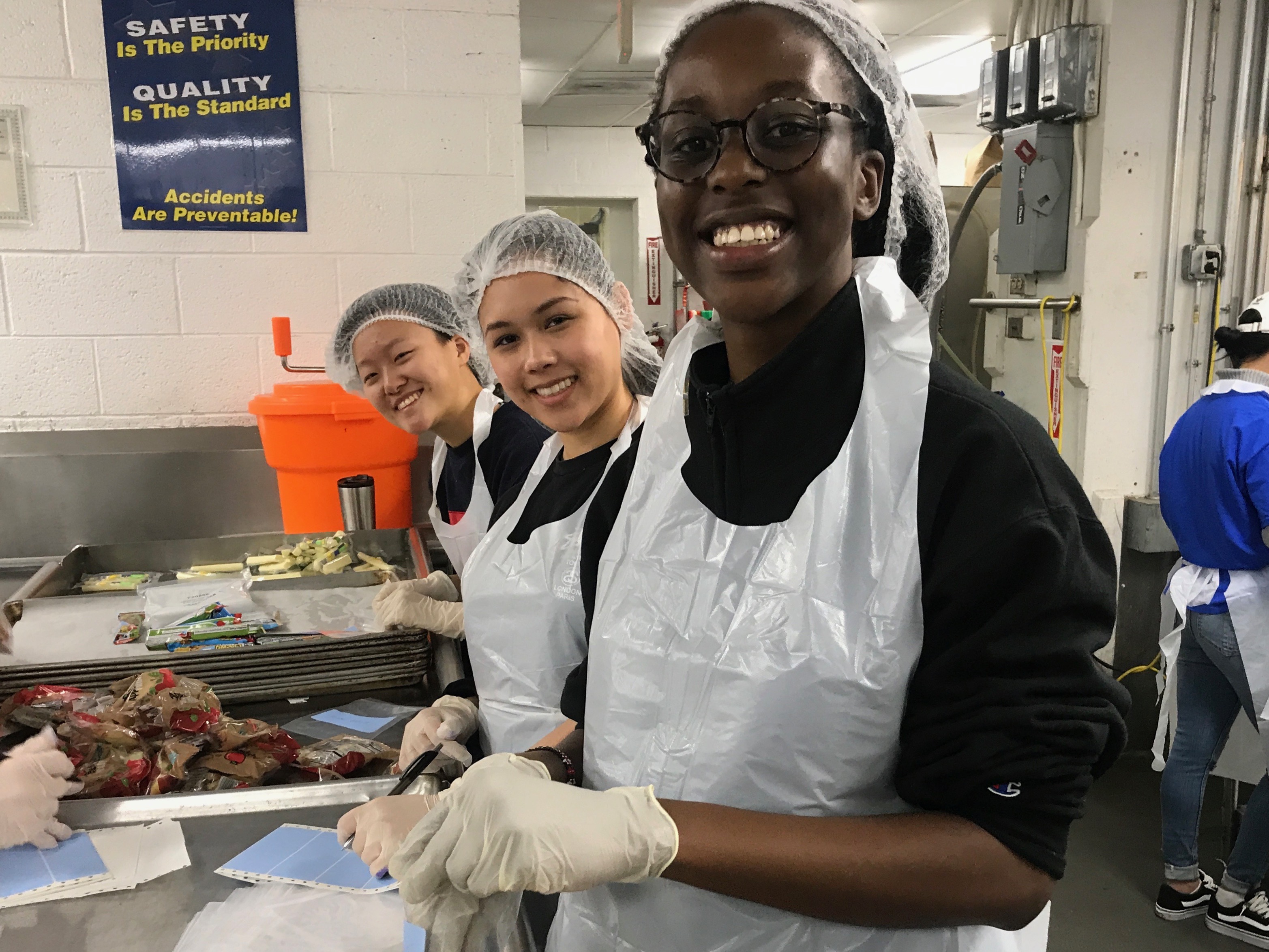 Three students wearing hairnet and aprons and gloves in a kitchen. 