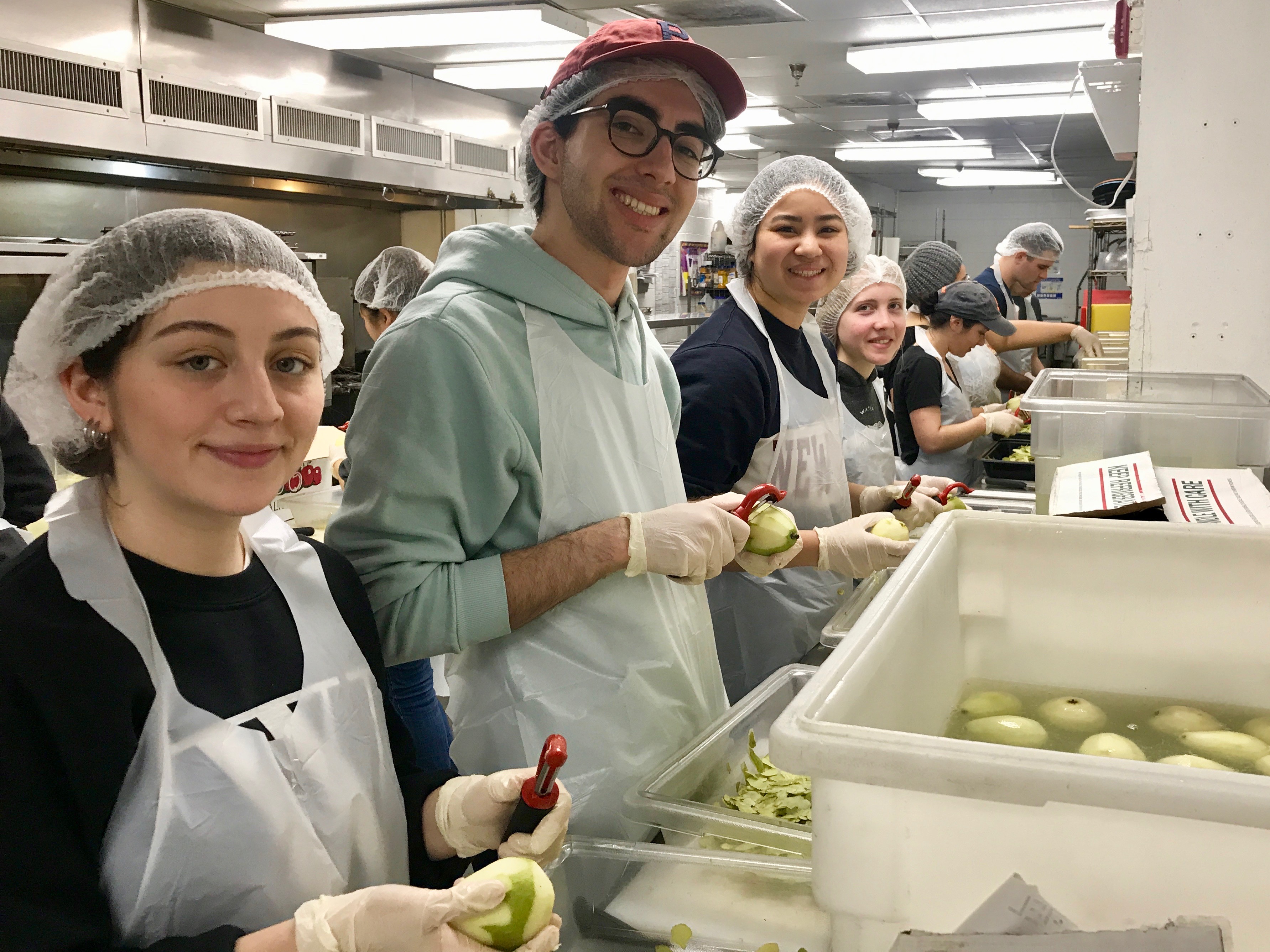 Four students with hairnets and aprons and gloves peeling apples in a kitchen.