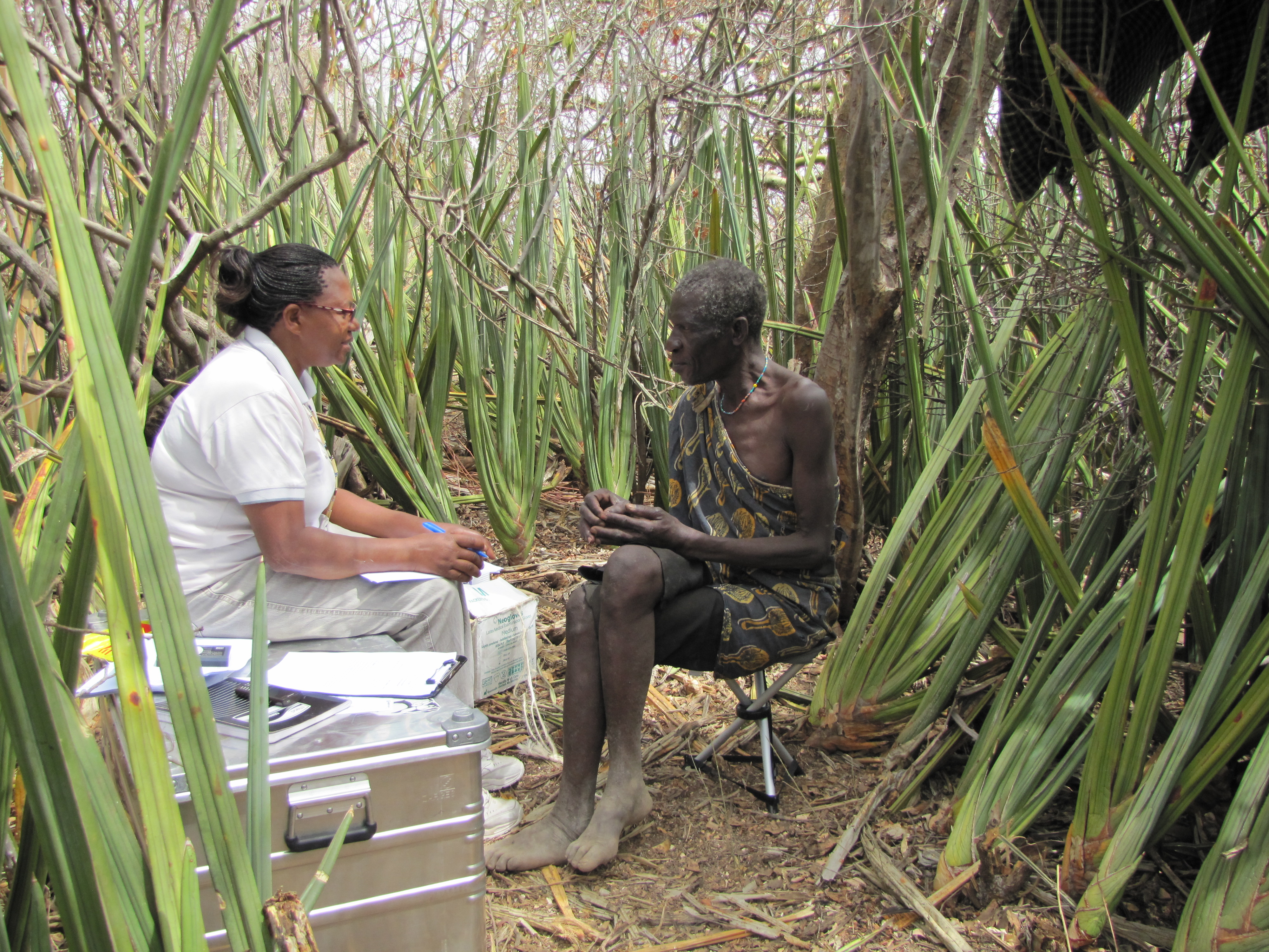 Person in white nurses outfit sits opposite person in traditional dress amid thick vegetation