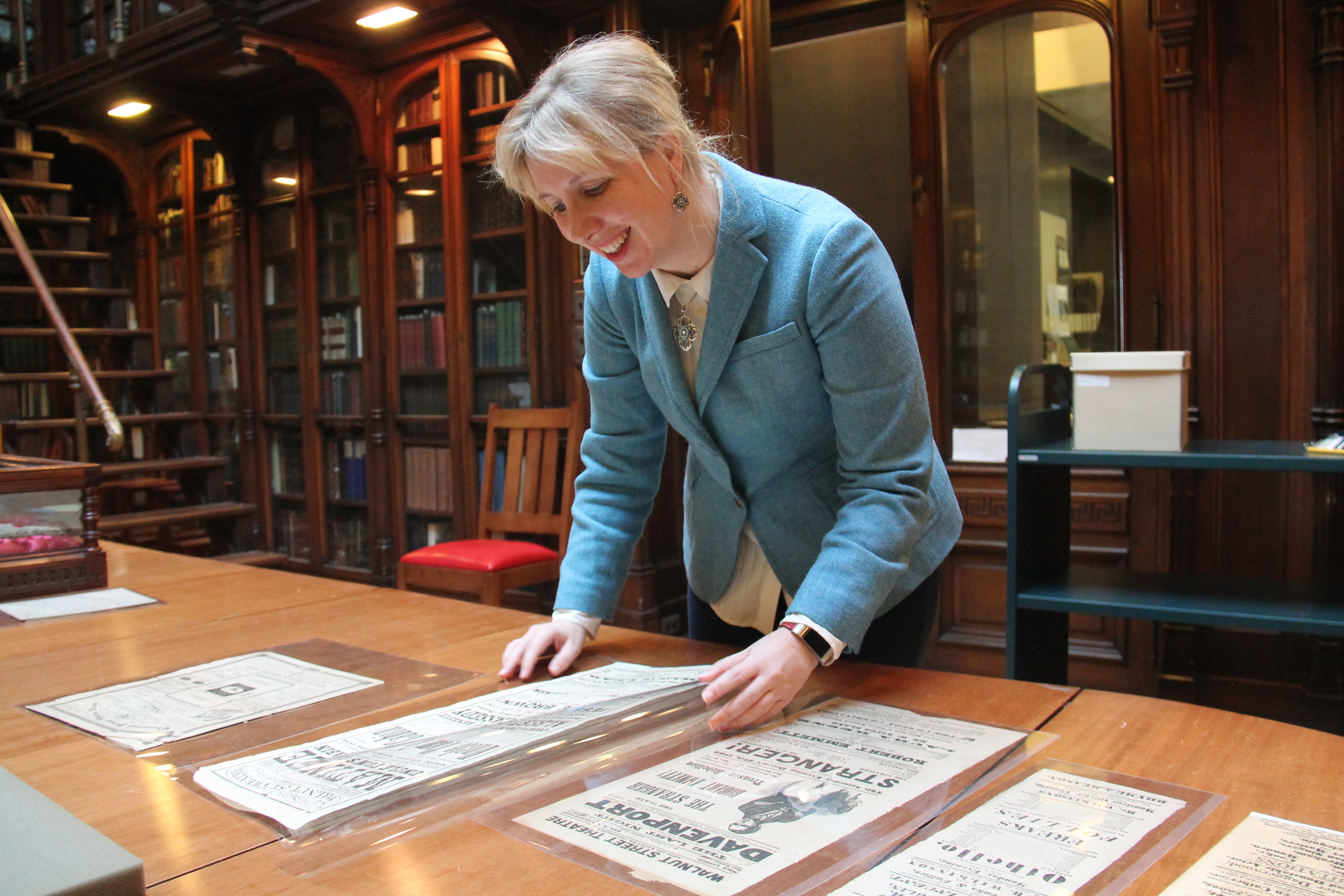 Librarian examines several playbills spread out on wood conference table in a historic library room lined with bookshelves filled with books.