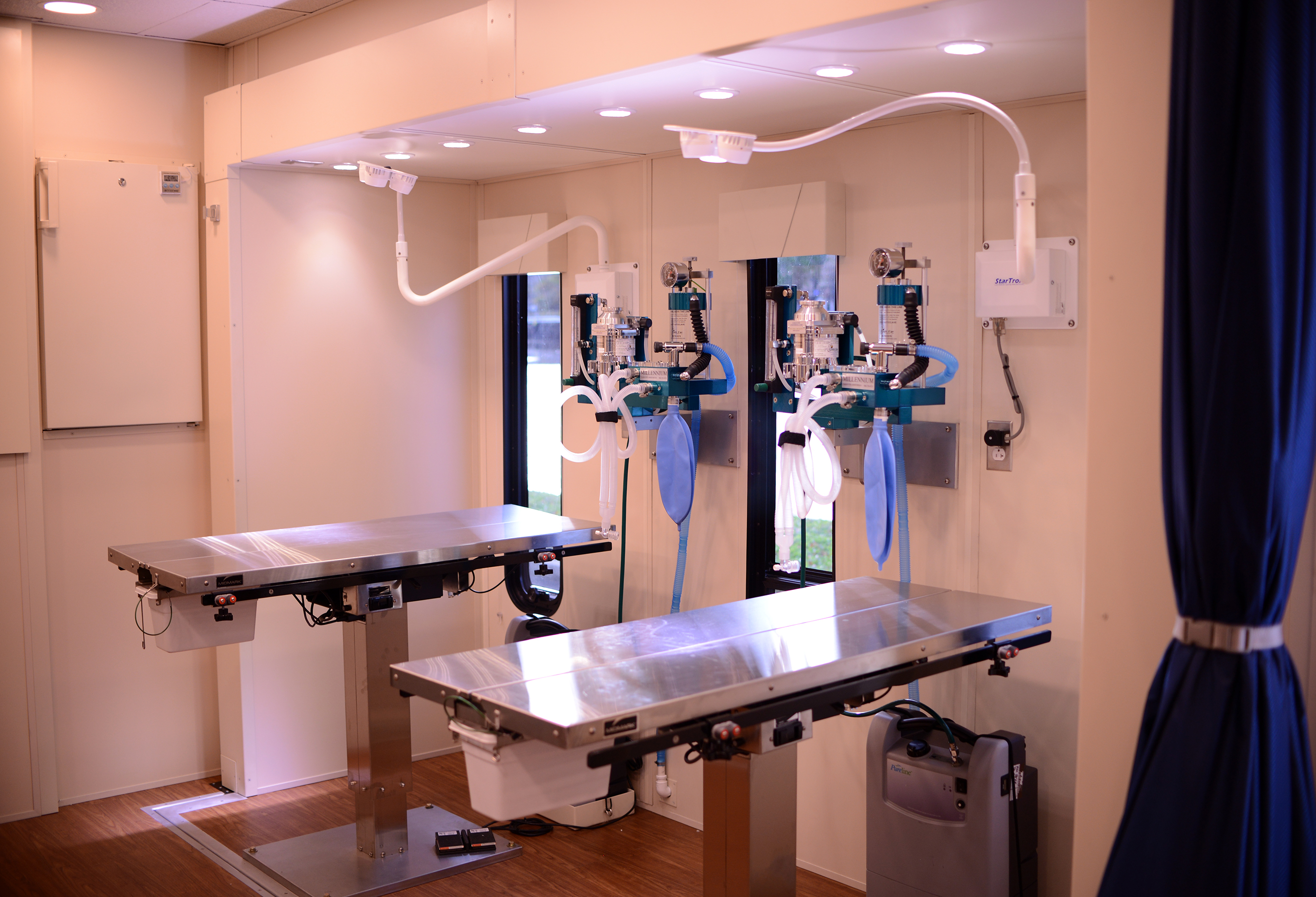 Two metal surgical tables with other surgical equipment above are shown inside a clinic space